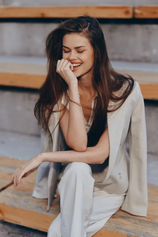 Young woman smiling confidently in beige business suit sitting outdoors on wooden steps, bright natural light, casual fashion, positive emotion, spring season