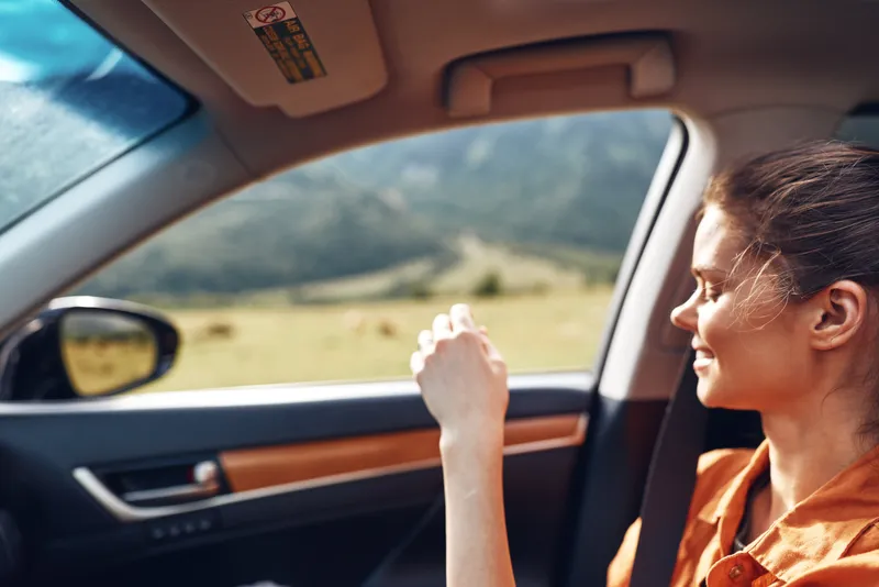 Woman in a car looking out the window during a road trip, enjoying scenic landscape with warm sunlight, relaxed expression and travel vibe in a modern vehicle.