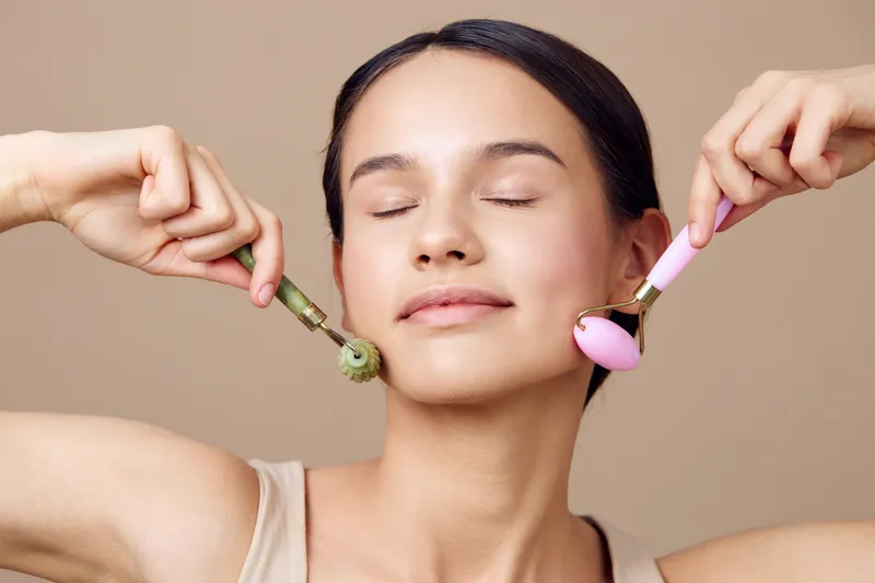 Young woman enjoying facial massage with jade and rose quartz rollers, promoting relaxation and beauty, against a neutral backdrop