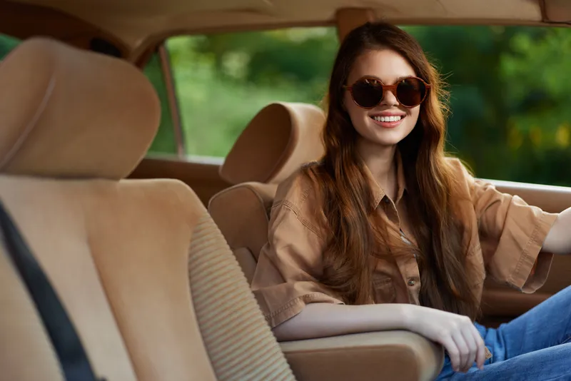Smiling young woman with long hair wearing sunglasses in a vintage car interior, embodying joy and freedom in a warm, sunny atmosphere.