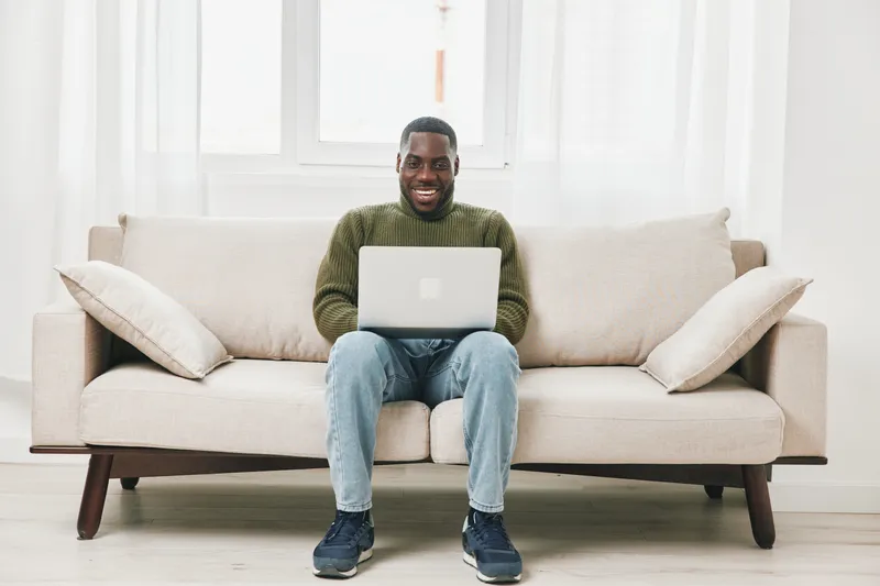 Happy black man working on a laptop at home on a cozy sofa, wearing a green sweater and jeans, smiling in a bright and airy living room setting