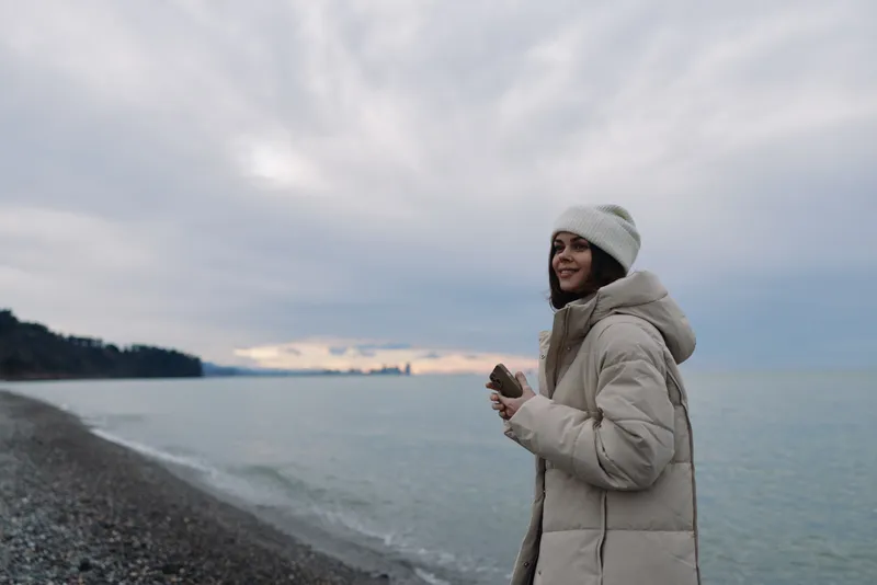 Winter woman in a coat at a cold beach by the sea, serene portrait in muted light, breeze in hair, cloudy sky, calm horizon, outdoor coastal mood.