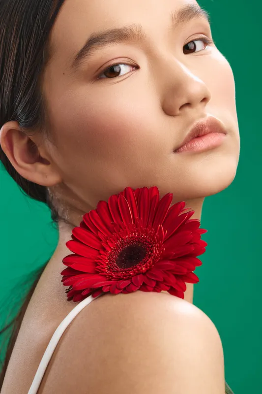 asian woman poses in a studio portrait with a bold red flower at her neck, soft lighting, and a vibrant green backdrop, exuding elegance and poised confidence.