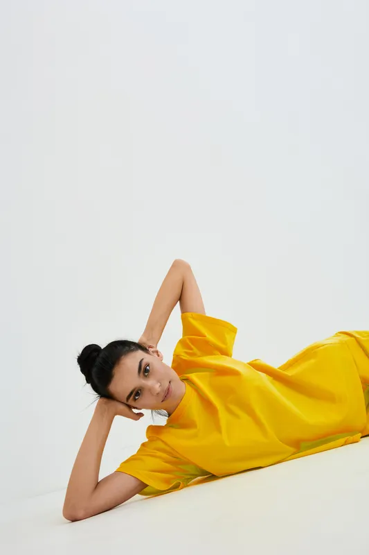 Young woman lying down wearing a bright yellow shirt, posing thoughtfully against a clean white background in a minimalistic style.