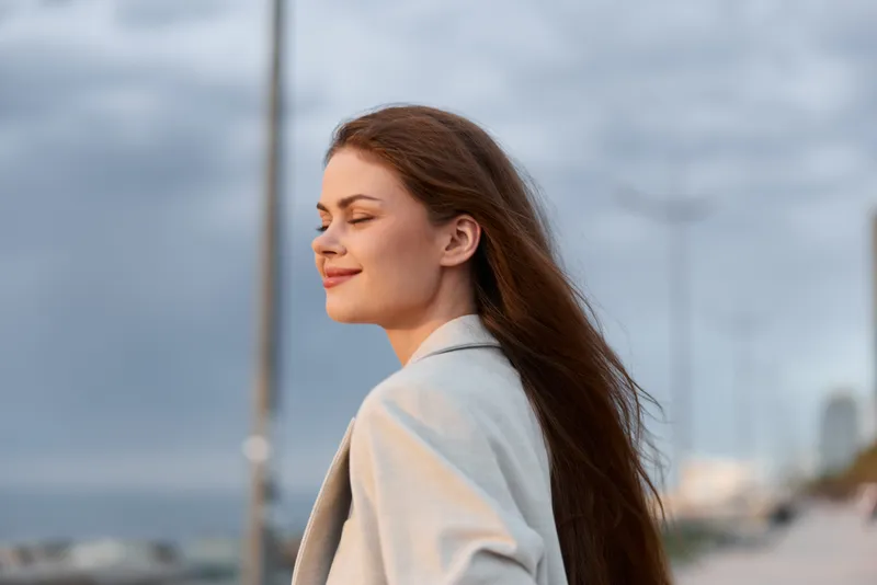 Woman with long hair in light coat enjoying peaceful outdoor moment by sea with cloudy sky, calm expression and natural wind background