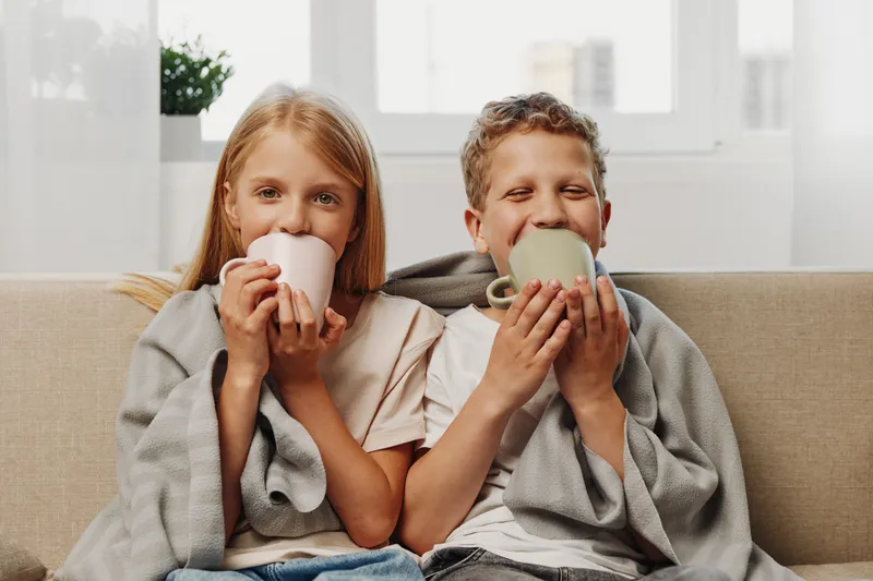 Two children wrapped in blankets enjoy warm drinks on a cozy sofa in a bright living room. Concept for comfort, childhood, warmth, and family time.