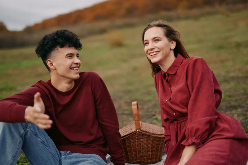 Two friends share a candid picnic moment in a meadow with a wicker basket, their authentic smiles and relaxed posture conveying credibility, genuine laughter and warm everyday companionship.