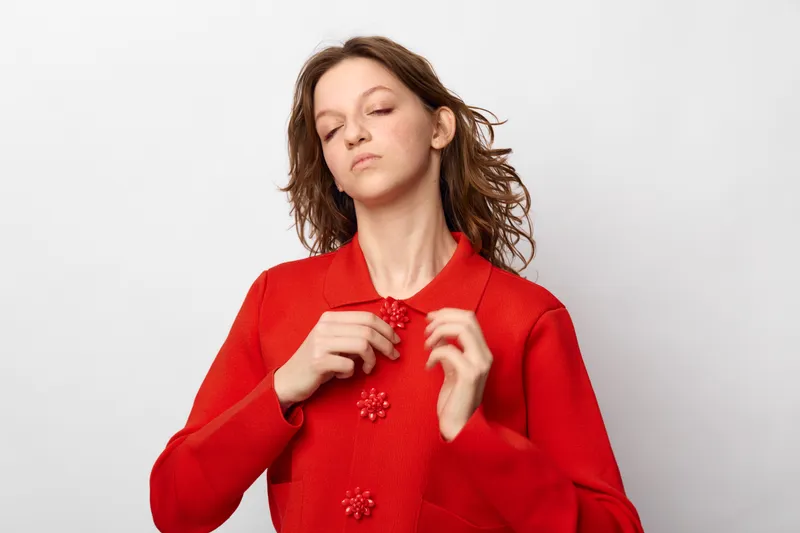 Young woman in red sweater buttoning up clothes with closed eyes. Isolated on white background. Concept of fashion, casual style, and self-confidence in studio lighting.