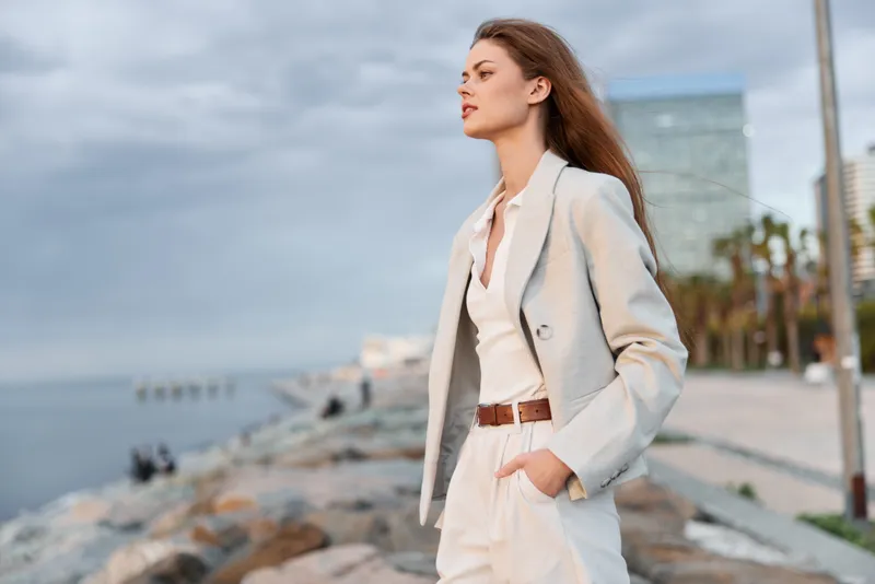 Confident woman in business attire walking by the water, showcasing elegance and professionalism against a modern urban background, perfect for corporate themes.