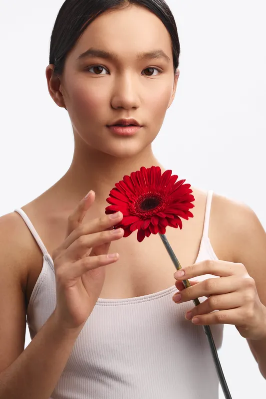 asian woman holds a red flower in a studio portrait, serene expression and soft lighting. The minimalist setup highlights grace and focus, conveying calm elegance.