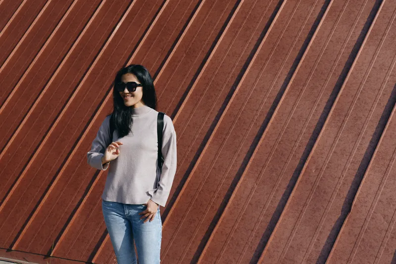 Woman wearing sunglasses, casual gray sweater and jeans, standing against a diagonal rust wall for urban fashion, outdoor street style, relaxed pose, contemporary photographic composition, moody