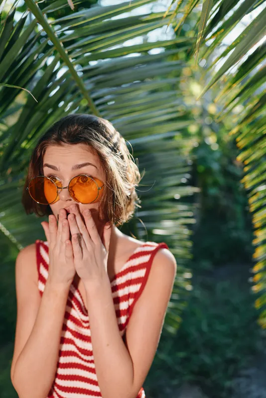 A cheerful young woman wears orange sunglasses and a striped red and white top, standing among tropical palm leaves in bright sunlight, creating a fun, summery vibe.