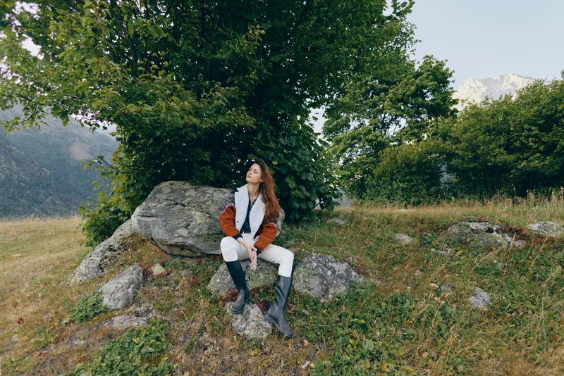 Woman sitting on rock in mountain meadow under tree, wearing jacket and boots, relaxed outdoors portrait enjoying nature and scenic landscape with distant peaks and green grass.