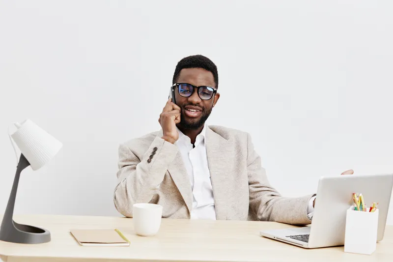 Confident young man of African descent with curly hair and beard, wearing glasses and beige blazer, sitting at modern office desk with laptop and stationery, smiling and talking on phone, light gray b