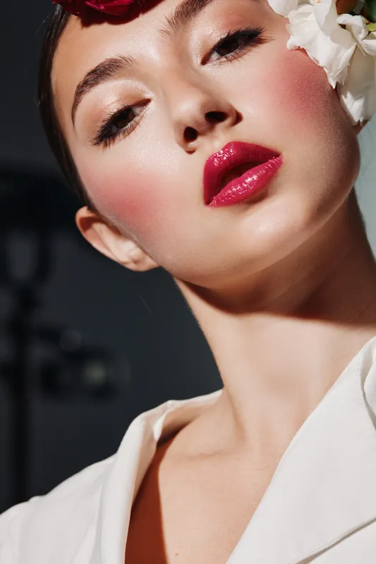 Close-up of a young woman with glossy red lipstick and flawless makeup posing against a dark background. She wears a white outfit and flower adorns her head creating fashion, beauty concept.