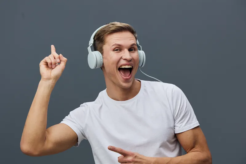 Smiling young man with headphones enjoying music, casual white t-shirt, expressive face, pointing up with index finger, gray background, studio shot, joyful mood, entertainment concept