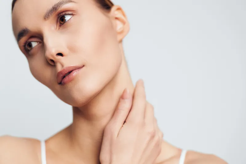 Close up portrait of a young brunette woman with a calm expression touching her neck in a studio setting, featuring soft lighting and a neutral background