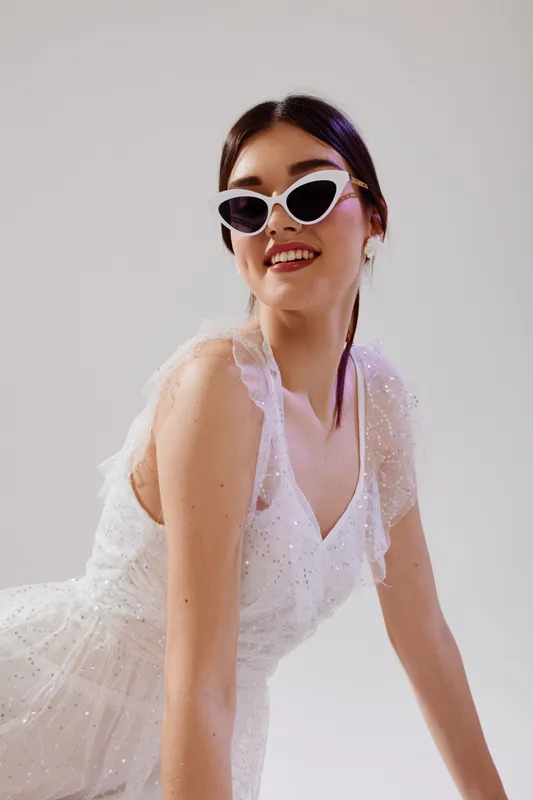 Young woman in a white dress and stylish sunglasses smiles confidently. Studio shot on a light background. Concept of beauty, fashion, and elegance.
