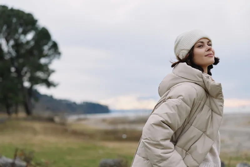 Woman wearing a puffy coat and knit beanie stands by a windy shoreline with trees and a distant horizon, capturing a calm outdoor moment of winter fashion and nature photography by the sea