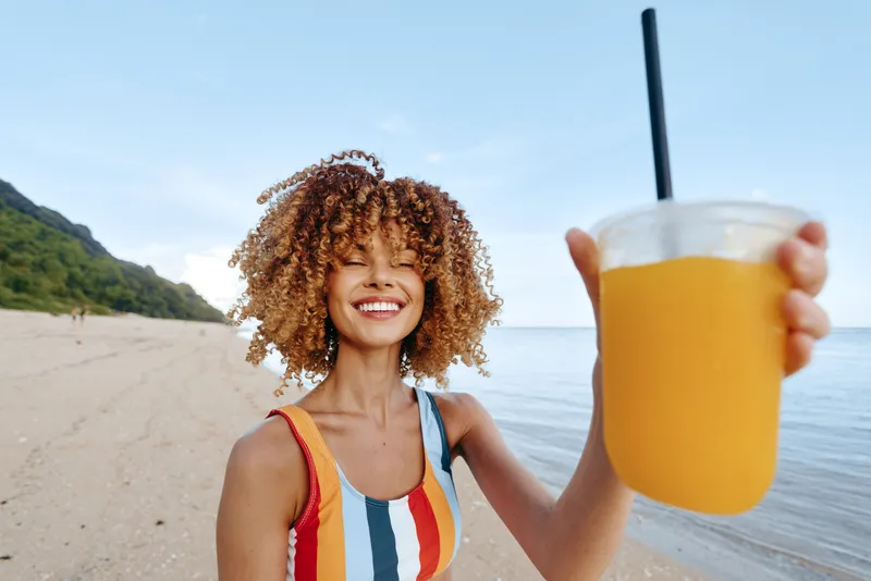 Cheerful woman on a sunny beach holds a bright drink toward the camera, smiling with eyes closed, wind in hair, colorful swimsuit, relaxed summer moment by the ocean, light breeze and natural colors