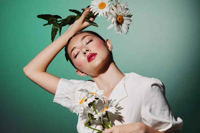 Young female model with bold red lipstick holds daisies while posing with closed eyes against green background. Creative fashion and beauty portrait in soft studio light and airy mood.