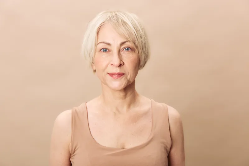 Portrait of a mature woman with short blonde hair wearing a beige tank top against a neutral background. Concept for aging, natural beauty, confidence, and wellness.