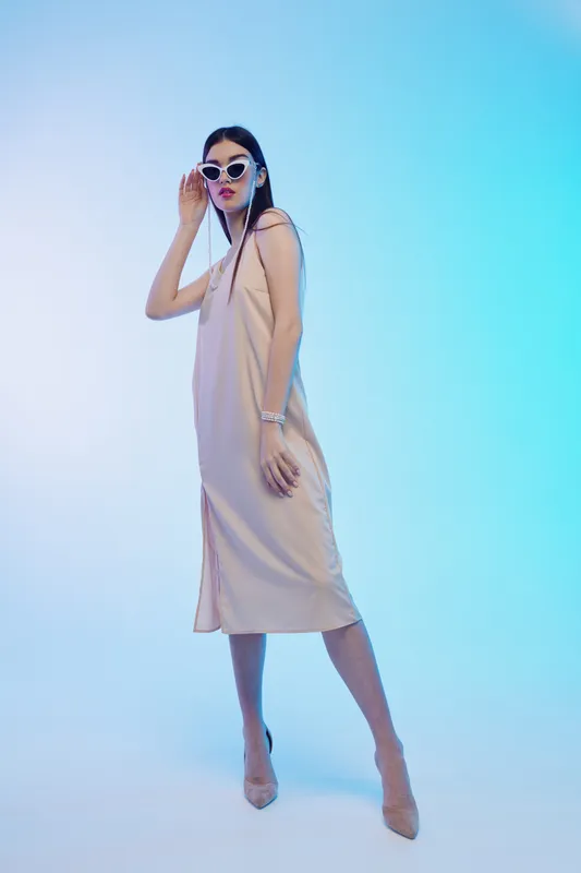 Woman beauty fashion model in beige dress and sunglasses poses confidently in studio with blue gradient background. Concept for style, elegance, and modern trends.