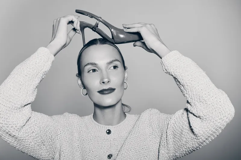 Young Caucasian woman with fair skin holds high heel shoes above her head, wearing knitted sweater and hoop earrings. Concept for fashion, style, and creativity.