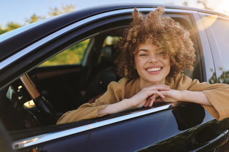 Smiling woman in a car, lifestyle concept with sunlit warmth and casual charm, candid portrait through an open window, conveying wellness and relaxed travel.