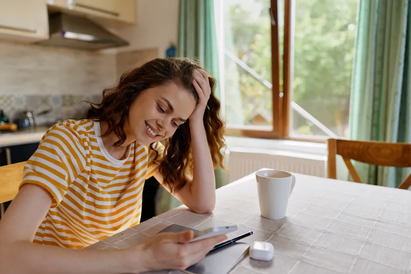 Woman in striped shirt sits at a table in a bright kitchen, using a tablet with a smile, capturing a casual home lifestyle moment bathed in natural daylight today.
