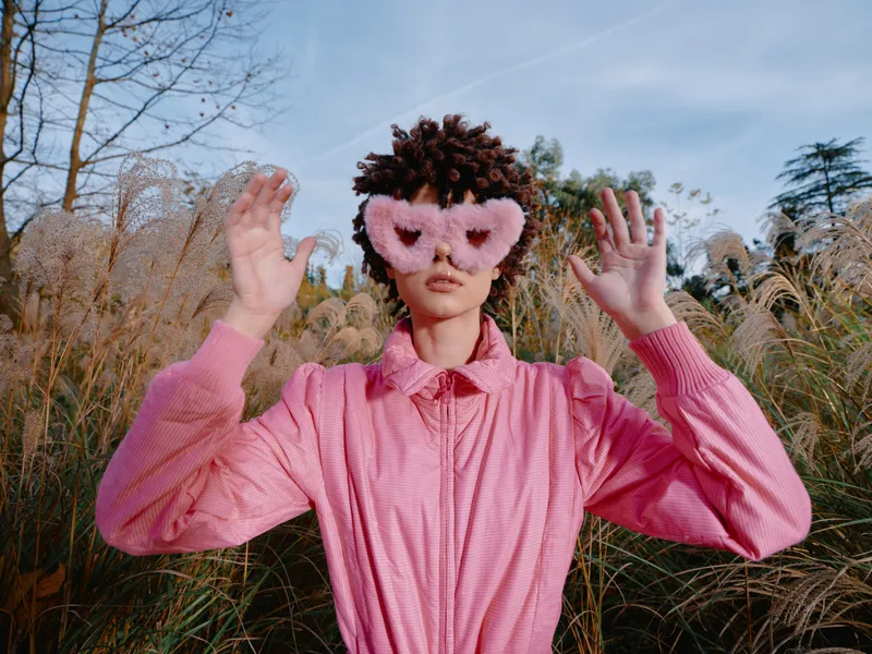 Whimsical person in a pink shirt wearing a heart fluffy mask in a sunny field, playful pose with hands near head, vibrant outdoor mood and imagination