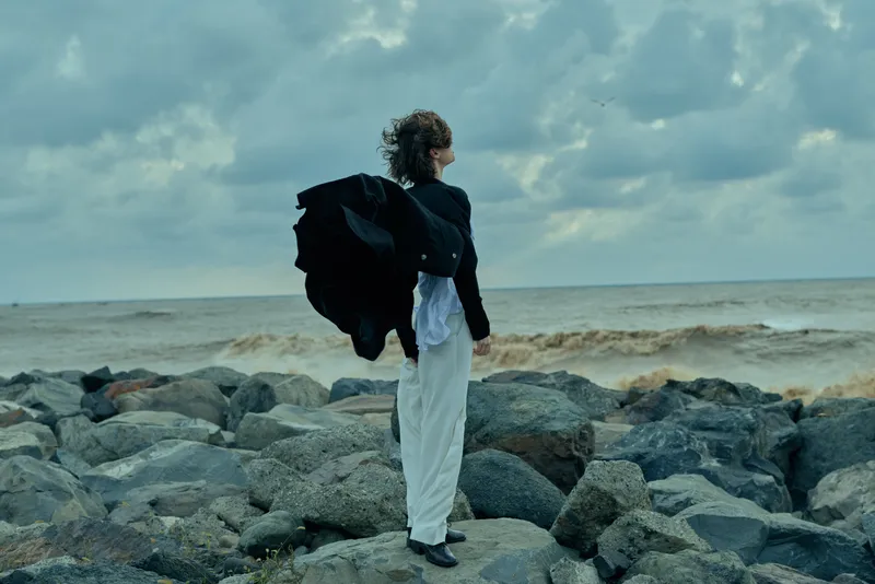 Wind swept figure on a rocky coast faces the sea under dramatic clouds, cloak billowing, showcasing resilience against the vast ocean.