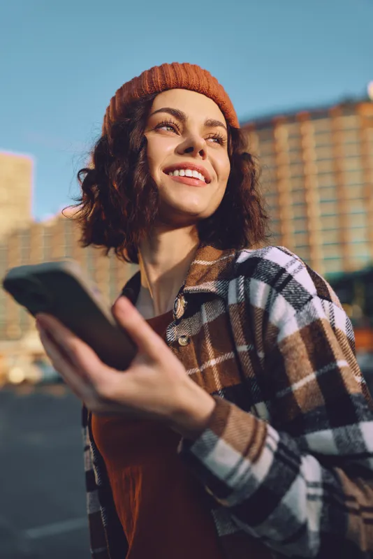 woman smile phone urban beanie plaid candid golden - A candid lifestyle portrait of a smiling woman holding a phone in city light, authenticity and golden hour glow, mindful living and emotional