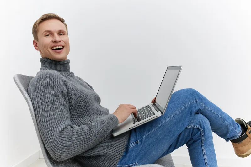 Smiling man working on laptop wearing grey sweater blue jeans sitting on chair isolated white background digital nomad concept