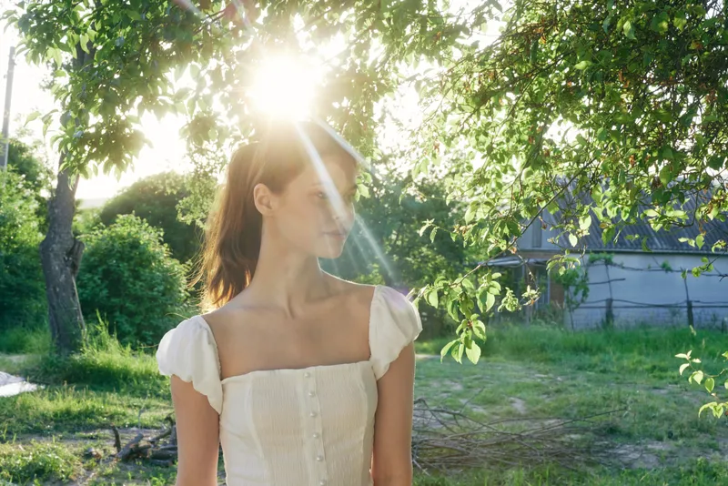 A young woman in a white dress stands in a sunlit garden, showcasing a reflective mood, surrounded by greenery and illuminated by warm sunlight