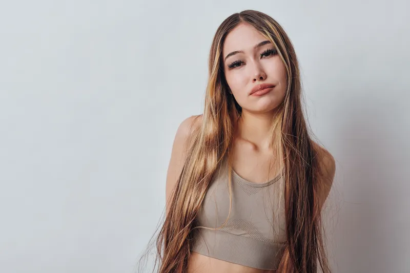 Young woman with long straight hair wearing a beige crop top standing against a neutral background looking at camera with a calm expression. Natural light and casual style.