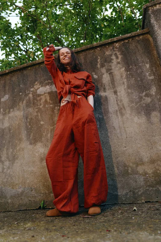 Smiling woman lifestyle outdoor fashion jumpsuit stands beside a weathered wall outdoors, showcasing confident candid style and bold color in a vibrant street moment
