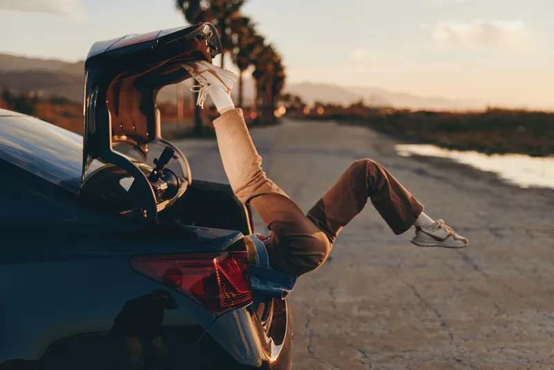 Woman lifestyle beside a car at sunset, seated on the road outdoors, boots and legs stretched, golden light on denim, quiet seaside evening, relaxed pose