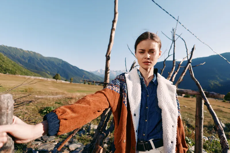 Woman portrait in mountains countryside by wooden fence wearing warm jacket and denim shirt, standing outdoors with fashion look and scenic valley landscape under clear sky.