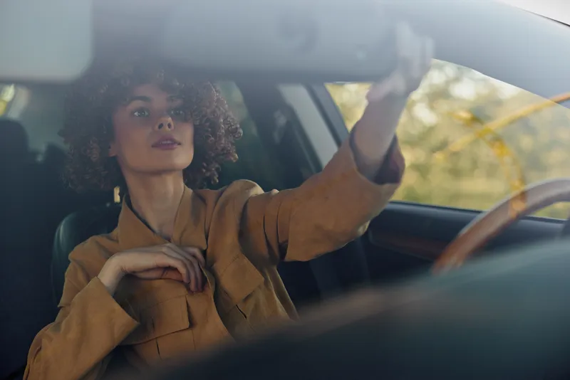 Woman drives a car in a casual retro style, interior shot featuring the steering wheel and window light, warm film color mood, confident expression, candid lifestyle portrait on the road