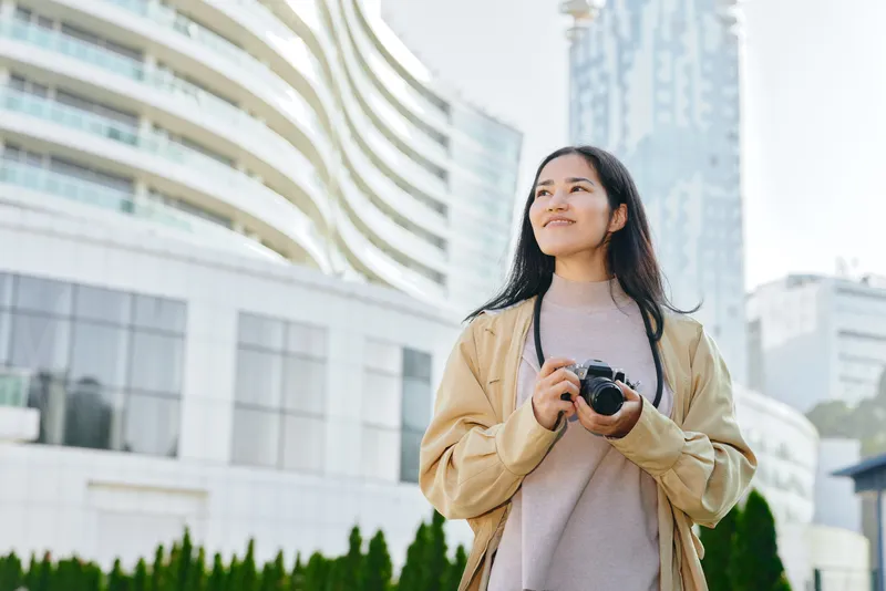 Young woman with a camera outdoors in a modern city, capturing urban street scenes and skyline with a relaxed pose, showcasing travel lifestyle and candid photography in daylight