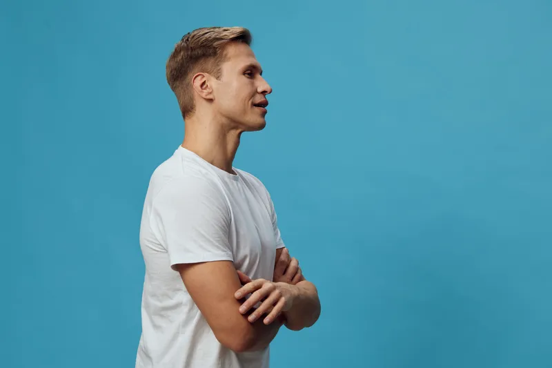 Confident young man with blond hair wearing casual white t-shirt standing sideways against a plain blue background studio portrait. People lifestyle concept