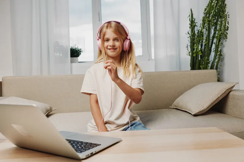 Smiling blonde girl with pink headphones enjoys music at home sitting on a beige sofa next to a laptop in a bright room with natural light and plants in the background.