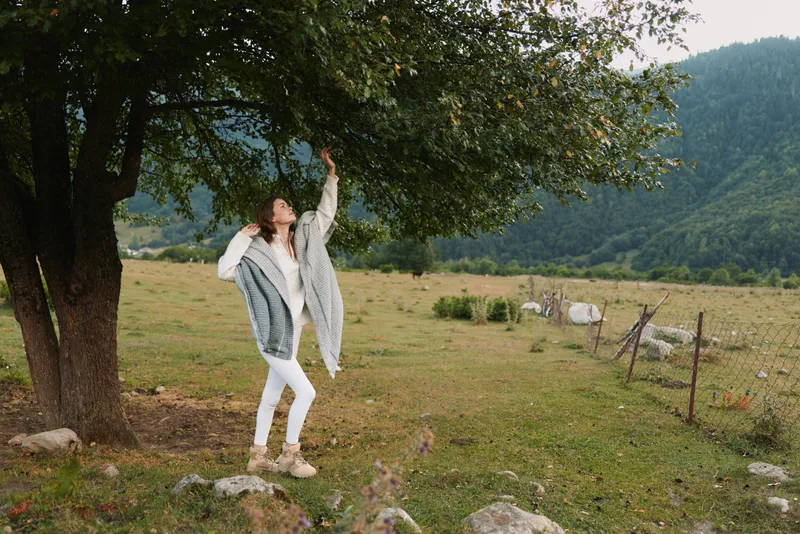 A woman in a light cardigan dances beside a tree in a rural field, capturing a carefree outdoor moment and a sense of freedom, a lifestyle scene of wellness and nature.