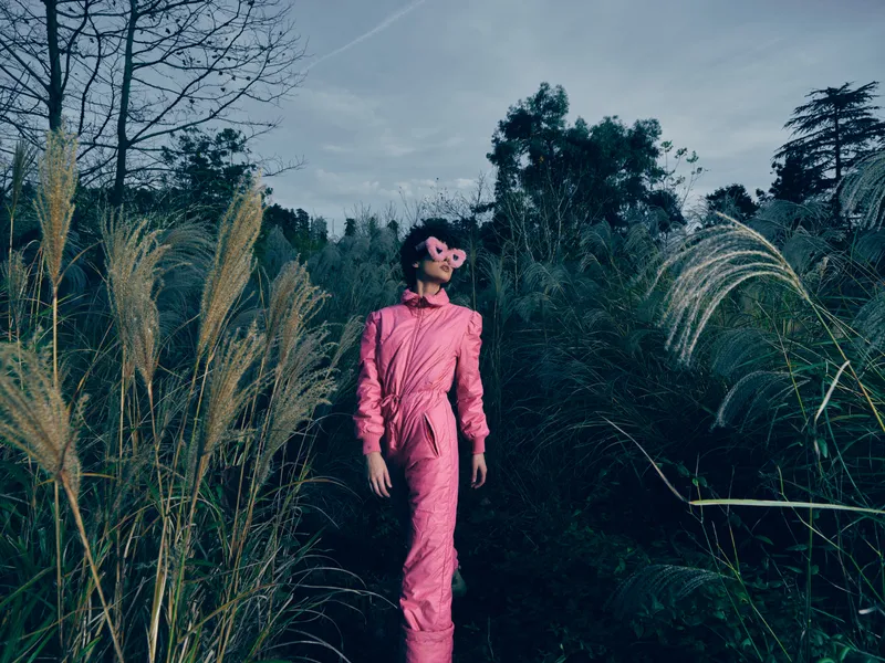Fashion model in a pink jumpsuit stands among tall grasses in a moody outdoor setting, showcasing bold color, confident pose and editorial style in a natural environment, candid atmosphere