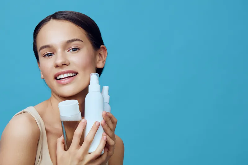 Happy young model holding skincare products against a blue background, showcasing a healthy skincare routine and glowing skin, perfect for wellness and beauty themes