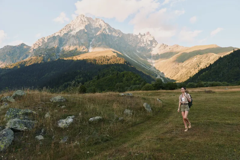 Hiking in a meadow with mountains in the distance, wearing a backpack and trekking gear. A scenic landscape captures outdoor lifestyle, nature, and calm adventure.