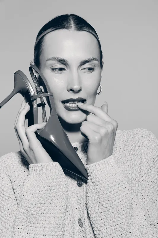White woman with pale skin holds high heel shoe near face biting finger in thoughtful pose. Fashion concept with stylish female model.
