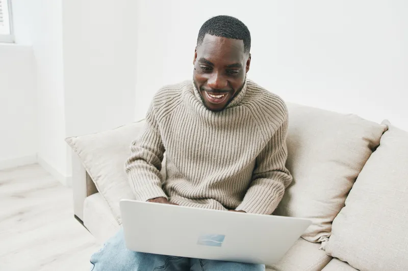 Smiling Black man working on a laptop at home, wearing a cozy beige sweater and sitting on a comfortable sofa, creating a relaxed and productive atmosphere