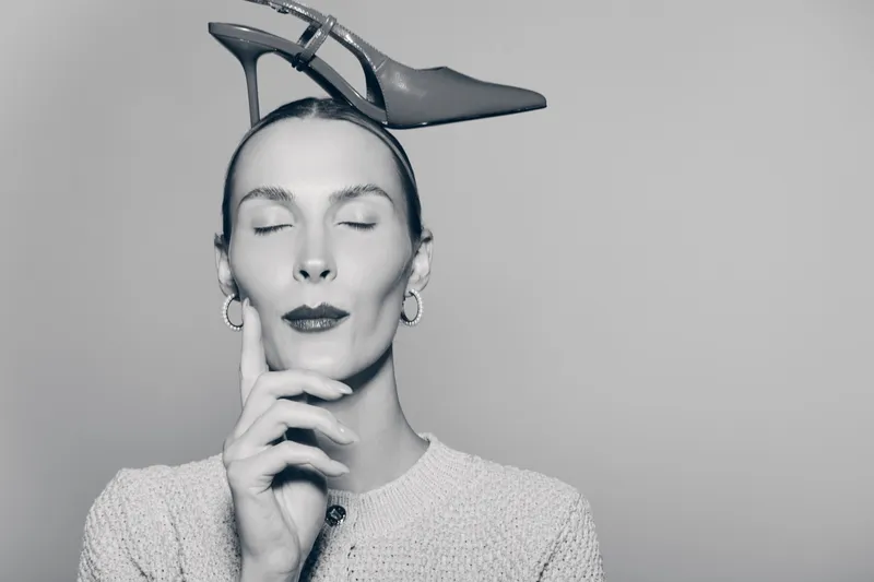 A white woman with pale skin and closed eyes balances a high heel shoe on her head, touching her face with fingers. Concept of fashion, surrealism, and creative portrait in studio.
